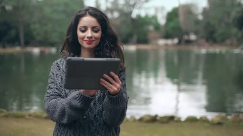 Smiling brunette using  the tablet at the park,lake in background Stock-Footage 74716058