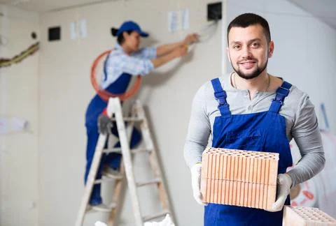 Smiling builder stacking red bricks on construction site indoors Stockfoto's