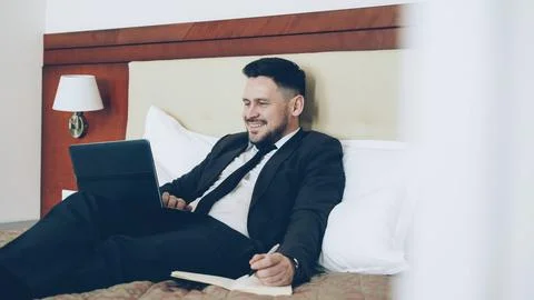 Smiling businessman lying on bed using laptop computer and writing notes at 스톡 사진
