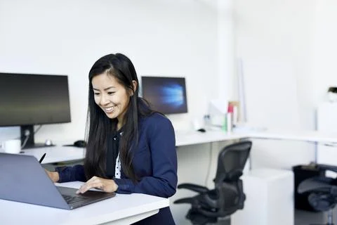 Smiling businesswoman using laptop computer on desk while standing against wall Stock Photos