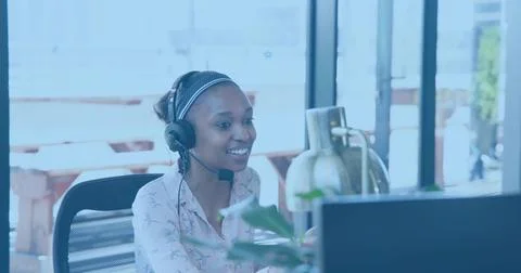 Smiling call agent in patterned blouse and headband, speaking on headset at desk 写真素材