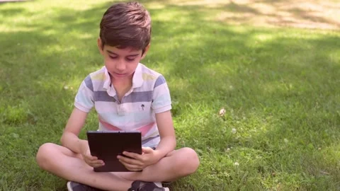 Smiling Caucasian little boy using Digital Tablet sitting on the grass outdoors Stock Footage 211403773