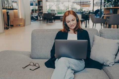 Smiling confident redhead lady using laptop and wireless headset, sitting on Stock Photos