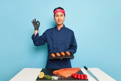 Smiling cook guy in blue chef's jacket holds black plate with sushi in his hand Stock Photos
