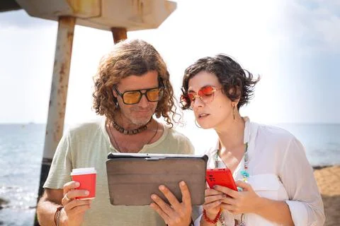 Smiling couple on the beach browsing digital tablet and smartphone apps. Stock Photos