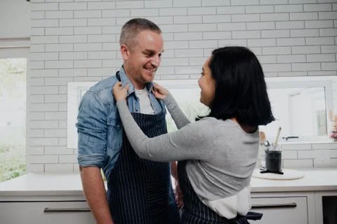 Smiling couple cooking in kitchen while wife puts apron on husband. Stock Photos