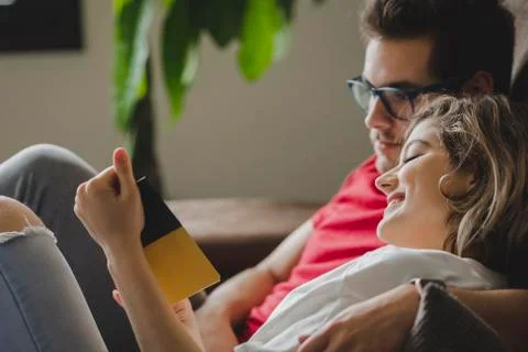 Smiling couple reading book while resting on sofa in living room at home Stock Photos