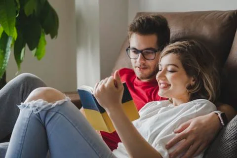 Smiling couple reading book while resting on sofa in living room at home Stock Photos