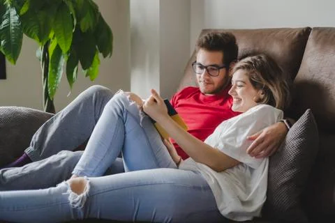 Smiling couple reading book while resting on sofa in living room at home Stock Photos