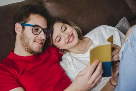 Smiling couple reading book while resting on sofa in living room at home Stock Photos