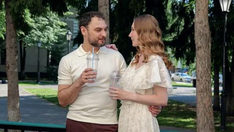 Smiling couple sharing cold drinks while walking through shaded city park on Fotos Stock
