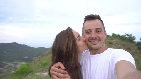 Smiling couple taking selfie in mountains, aerial city view. Woman kisses man Stock Footage 76510156