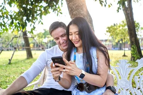 Smiling couple using the mobile while sitting on a bench outdoors during sunset Stock Photos