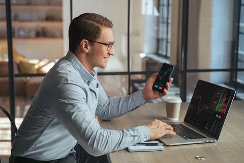 Smiling crypto trader with phone checking candlestick graph on laptop, buying Stock Photos