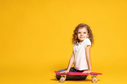 Smiling curly little girl in white polo, blue jeans, sitting with skateboard Stock Photos