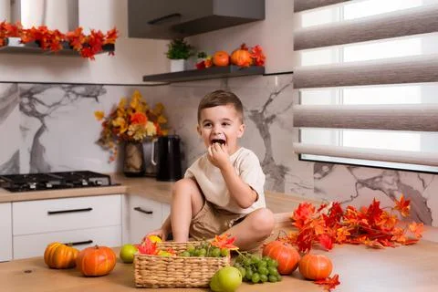 Smiling cute sitting on kitchen table with fruits, autumn leaves and pumpkin Stock Photos