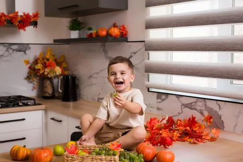 Smiling cute sitting on kitchen table with fruits, autumn leaves and pumpkin Stock Photos