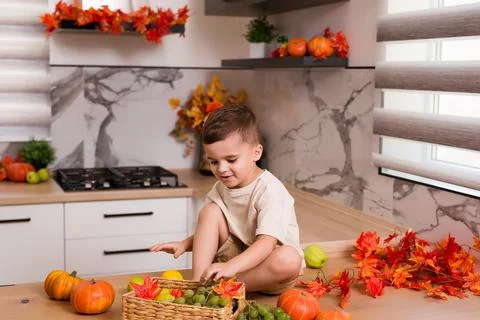 Smiling cute sitting on kitchen table with fruits, autumn leaves and pumpkin Stock Photos
