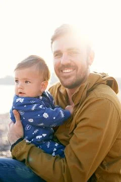 Smiling dad in a brown jacket sits and holds a small child in a blue overalls in Stock Photos