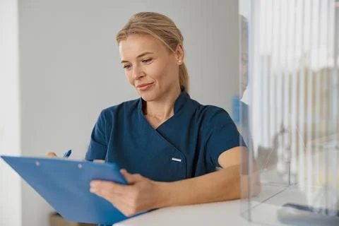 Smiling doctor making notes on clipboard standing near reception in clinic hall Stock Photos