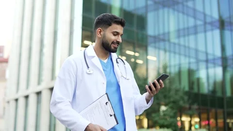 Smiling doctor in white coat using mobile phone while walking outside near  Stock Footage 315560335