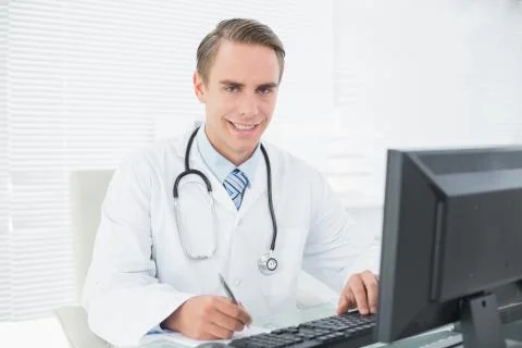 Smiling doctor writing note while using computer at medical office Foto stock