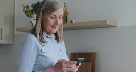 Smiling elder age woman using smartphone at home in modern apartment. 库存影片 334070909