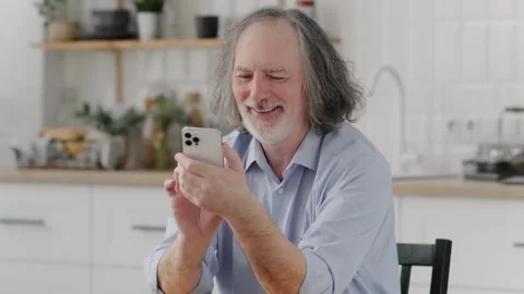 Smiling Elderly Man Using Smartphone While Sitting At Desk In Kitchen Interior Video stock 250162975