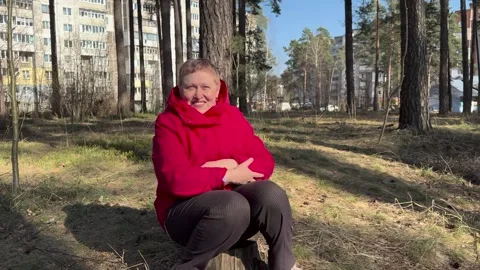 Smiling elderly woman in a red jacket sitting on a tree stump Vidéo 332420526