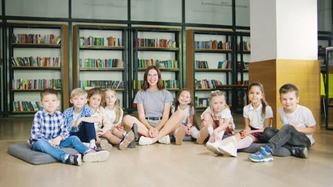Smiling elementary students sit with their teacher on cushions in a cozy school Video stock 284269536