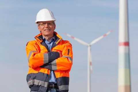 Smiling engineer with hard hat and protective clothing in front of wind turbi Stock Photos