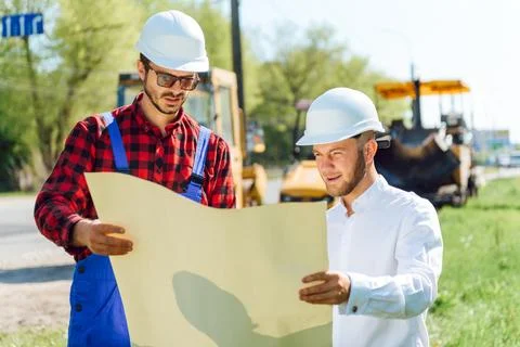 Smiling engineer with helmet standing in front of excavator on road constru.. Stock Photos