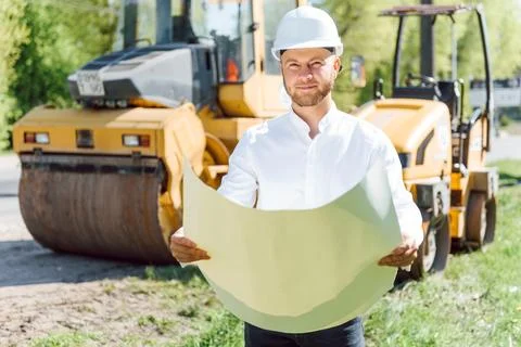 Smiling engineer with helmet standing in front of excavator on road constru.. Stock Photos
