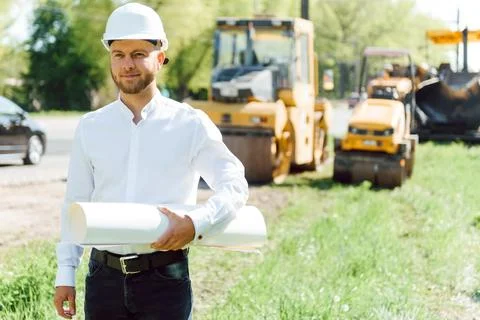 Smiling engineer with helmet standing in front of excavator on road constru.. Stock Photos