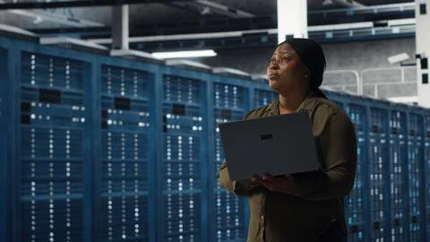Smiling engineer walking in data center with laptop, checking hardware Stock Photos