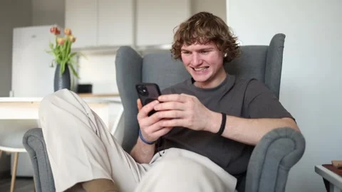 With a smiling expression, the young man sits in a chair holding his phone, like Stock Footage 302633791