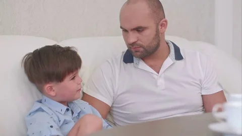 Smiling father talking to his son sitting on the white sofa and drinking tea Vídeos de archivo 76582716
