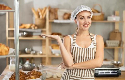 Smiling female baker gesturing invitingly in cozy bakery Stock Photos