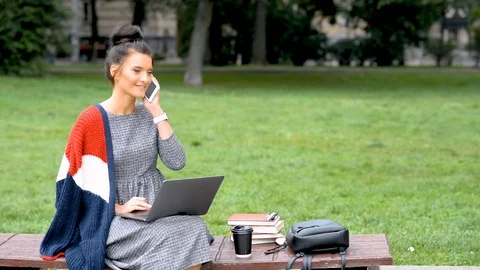 Smiling female student talks on cell phone while sitting with laptop in park. Stock Footage 117125585