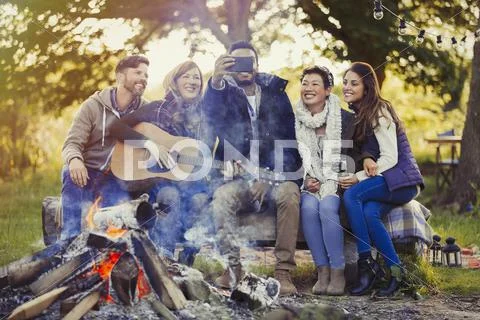 Smiling friends with camera phone taking selfie at campfire Stock Photos