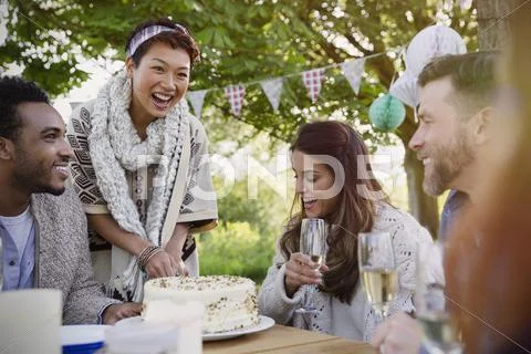 Smiling friends drinking champagne and cutting birthday cake at patio table Stock Photos