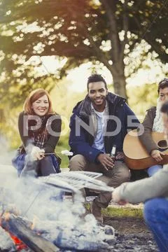 Smiling friends roasting marshmallows and drinking beer at campfire Stock Photos