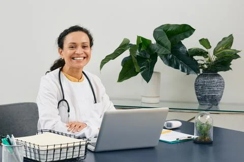 Smiling general practitioner sits at her workplace wearing a medical coat wit 写真素材