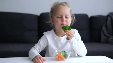 Smiling girl happily eats broccoli home Child puts green broccoli florets into Video stock 275529285
