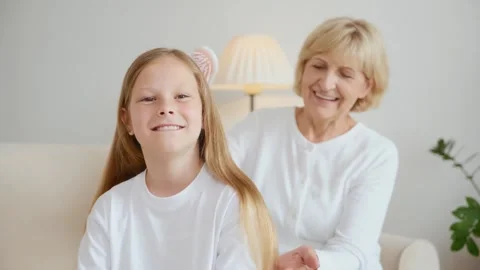 Smiling girl looks at camera while grandmother brushes her hair Video stock 315557554