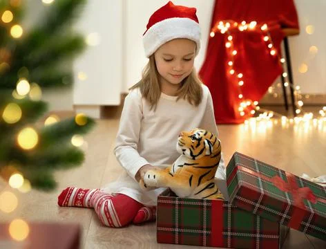 Smiling girl opens gift box with toy tiger next to christmas tree. Stock Photos