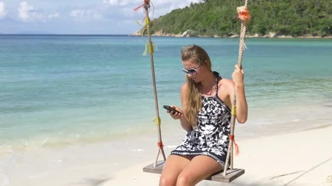 Smiling girl using phone on a swing at beach Stock Footage 74684609