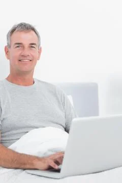 Smiling grey haired man using his laptop in bed Stock Photos