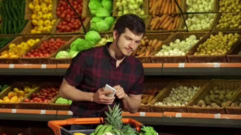 Smiling guy checks smartphone while shopping for fresh produce Stock-Footage 311947549