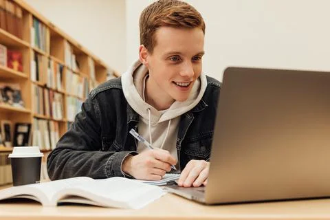 Smiling guy looking at laptop screen while sitting in library Stock Photos